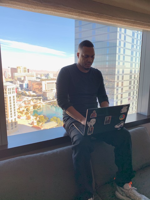  Freelance Software Developer male sitting on top of a couch in a Las Vegas hotel room, coding on his laptop 
        with a large window in the background over looking the Las Vegas stip.